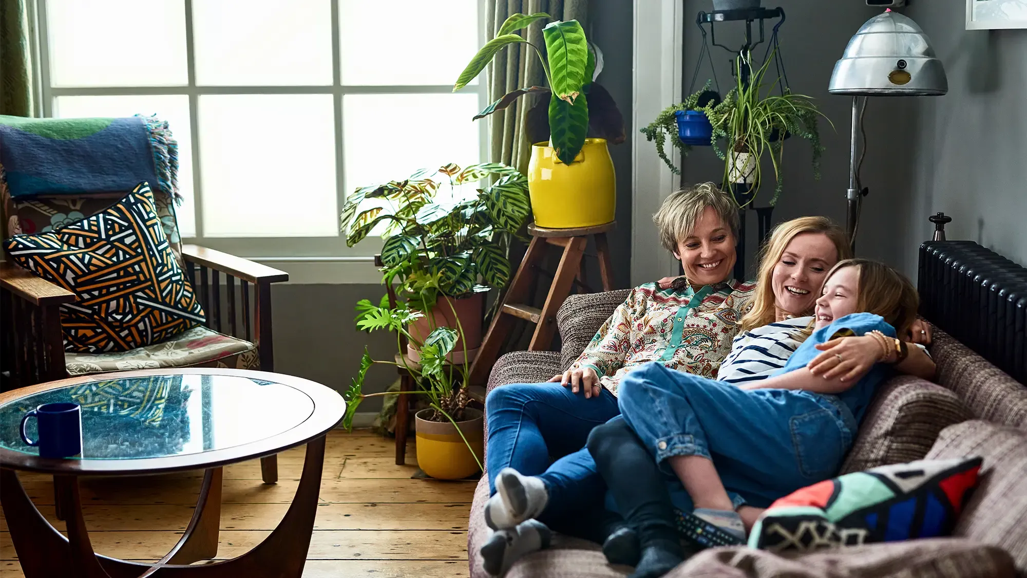 Three people are joyfully sitting on a couch in their new home. A child is cuddled between two adults, laughing. The room is decorated with vibrant plants and a yellow pot. The atmosphere is warm and cozy.