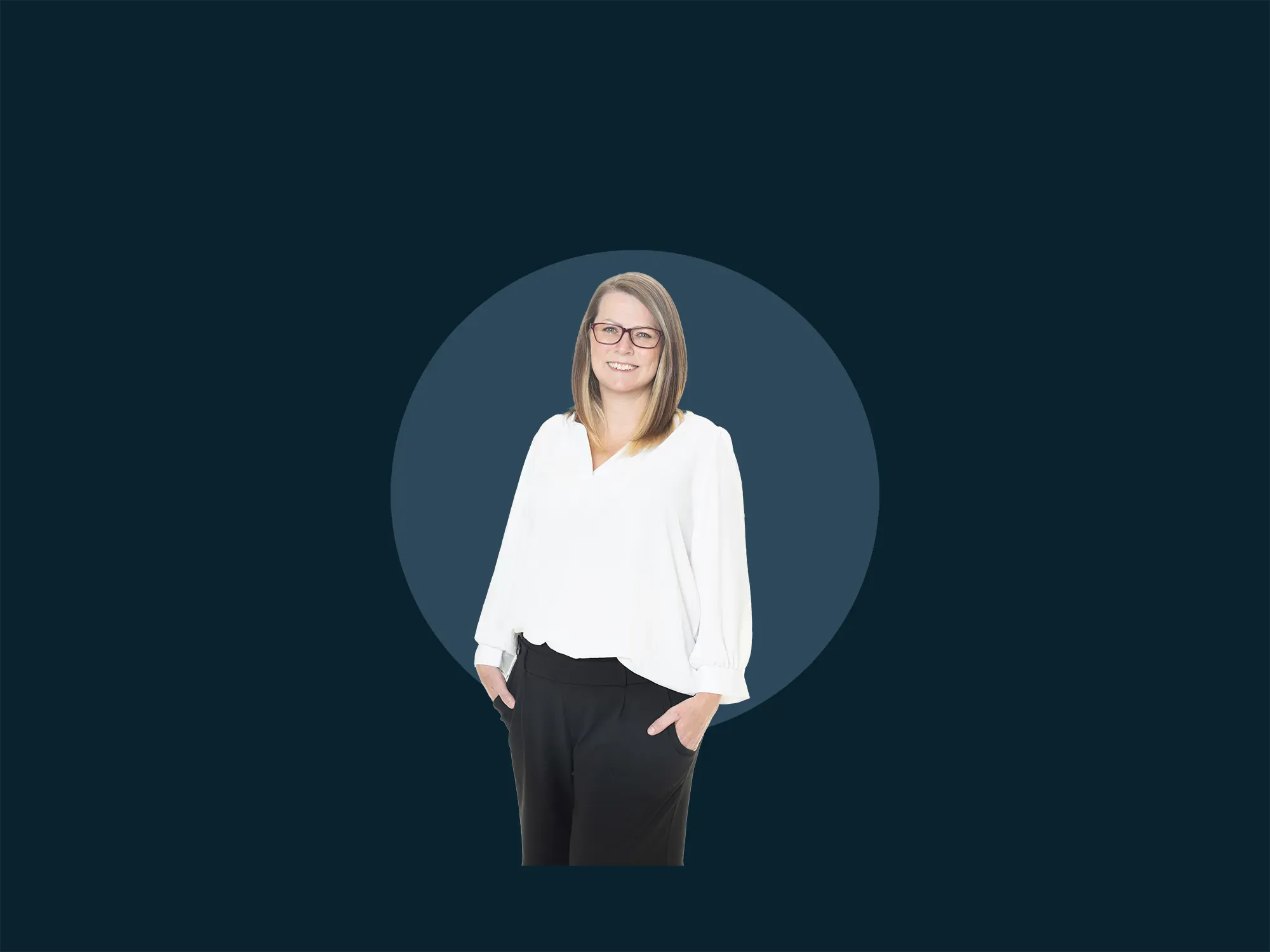 A woman in a white blouse and black pants stands smiling with one hand in her pocket against a dark blue background.