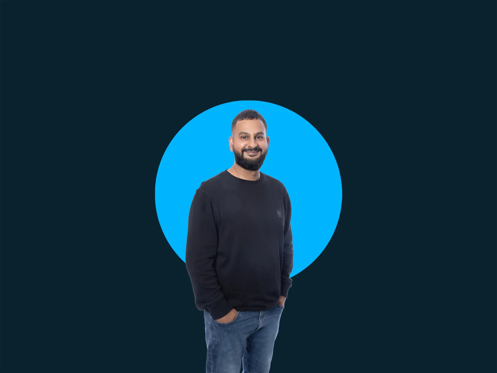 Smiling man in a black sweater and jeans stands against a blue circle backdrop on a dark background.