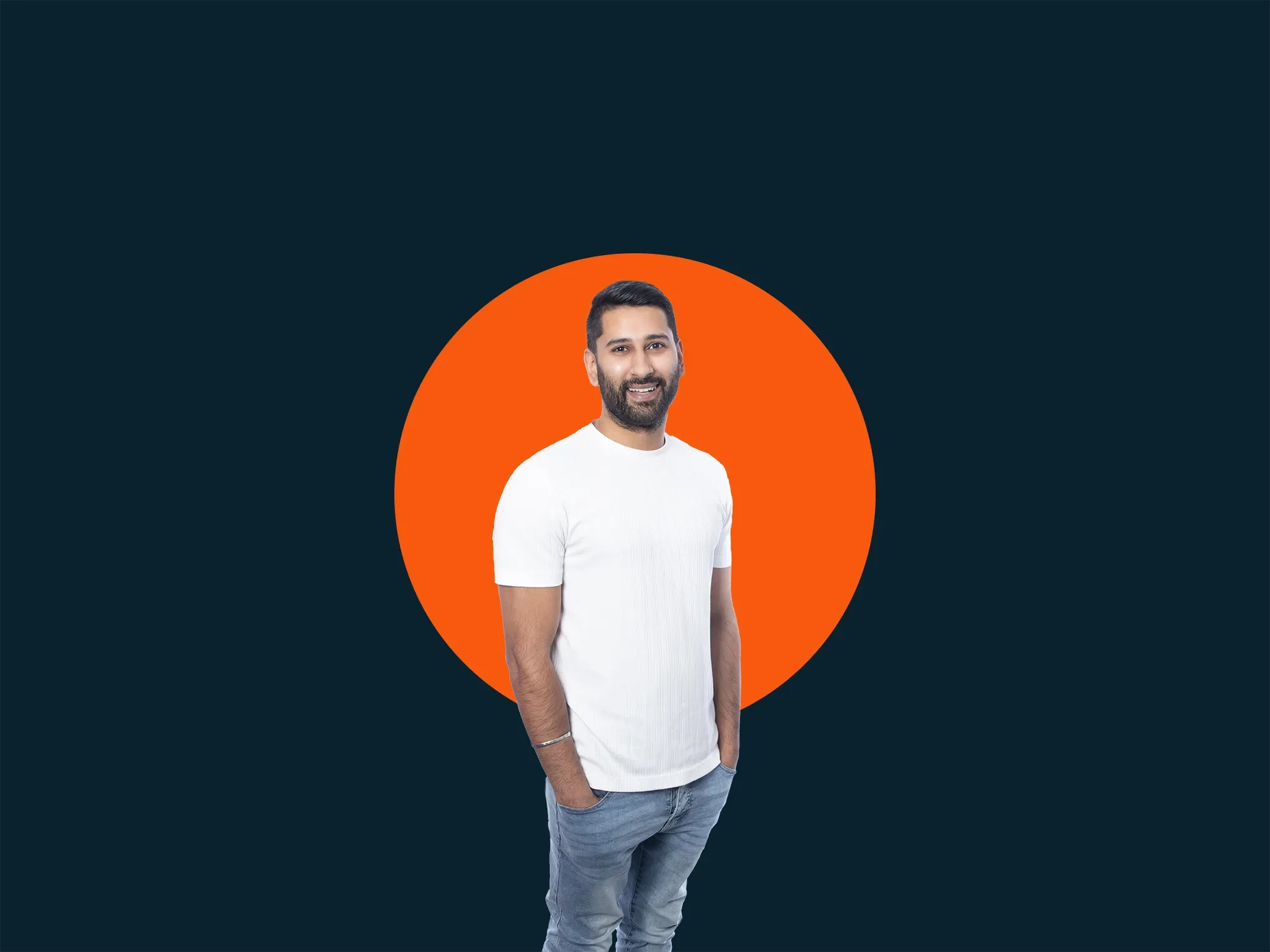 Man in a white T-shirt and jeans stands with hands in pockets, smiling, against a dark background with an orange circle behind him.