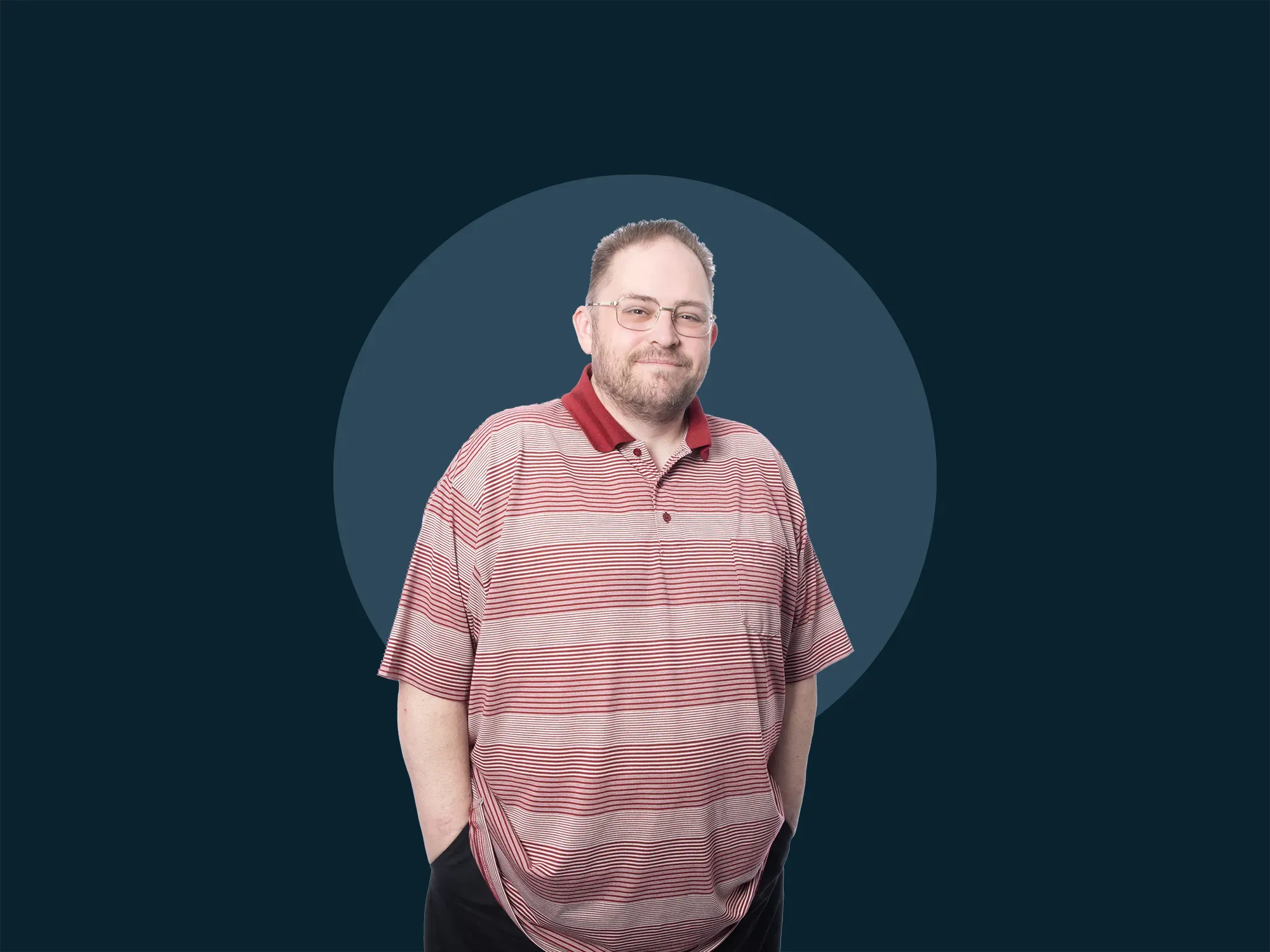 Sean Donald Deacon in glasses, wearing a red striped polo shirt, stands against a dark background with hands in pockets, subtly smiling.