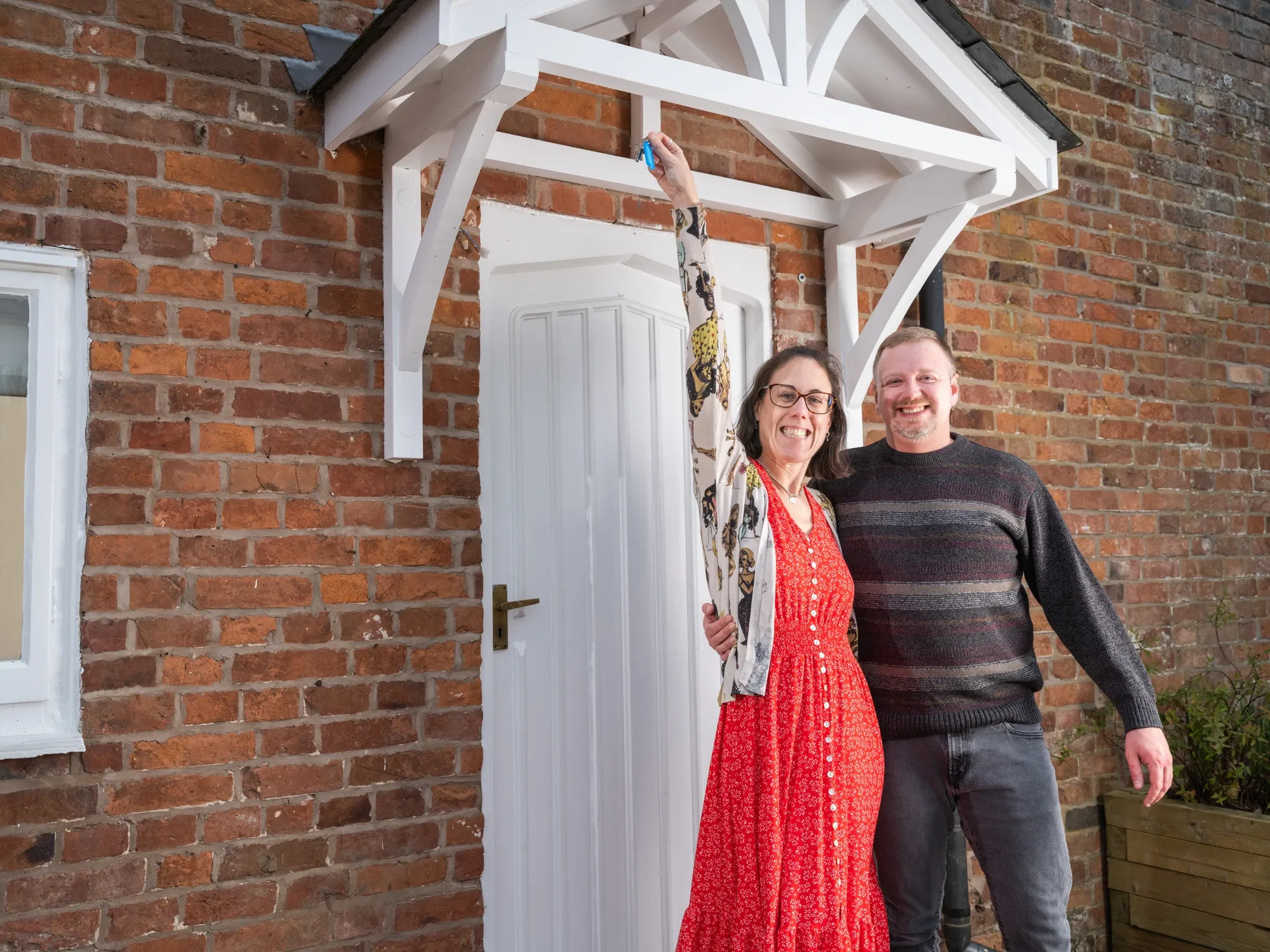 A smiling couple stands in front of a brick house, with the woman holding up a key, celebrating their new home bought with cash.