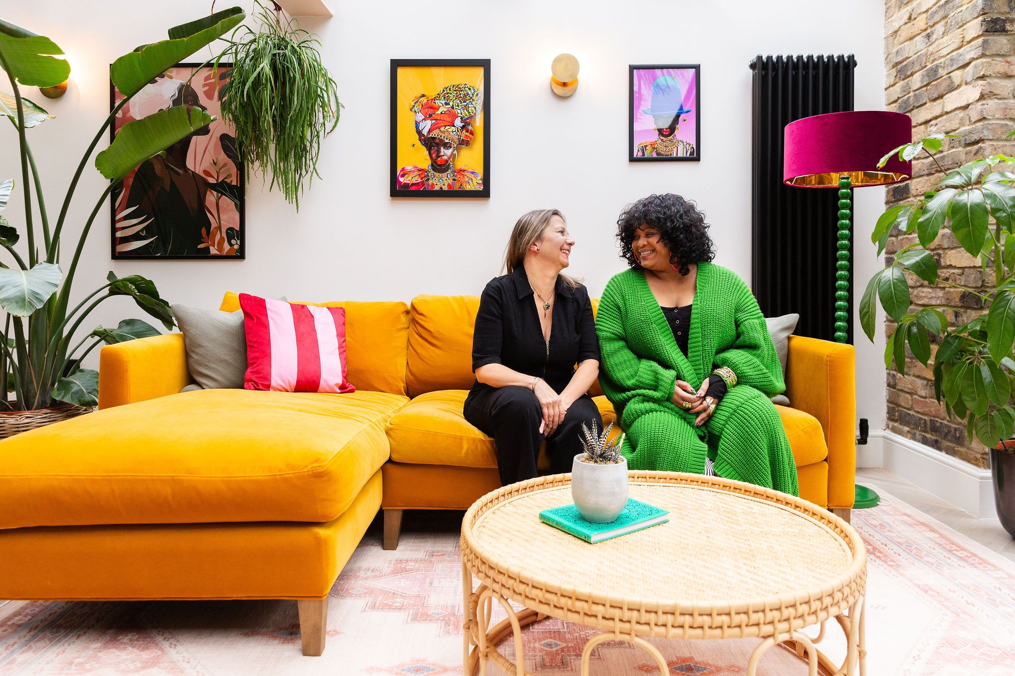 Two women sit smiling on a bright yellow sofa in a colorful living room with vibrant artwork and lush green plants.