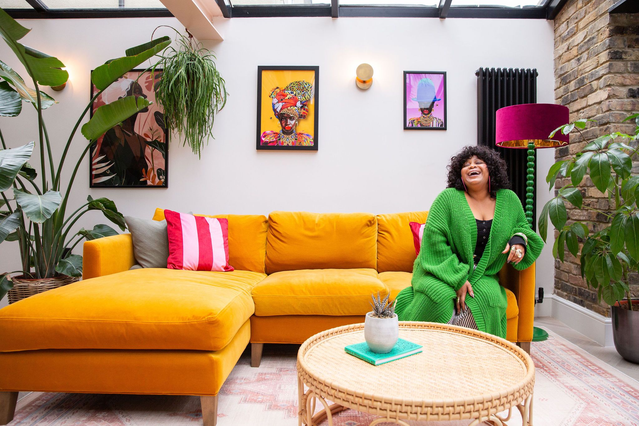 Woman in green sweater smiling on yellow couch in bright living room with plants and colorful wall art.