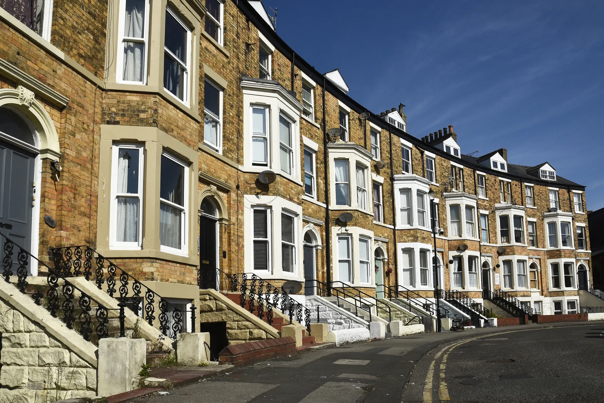 A row of Victorian terraced houses with brick facades and bay windows, featuring black wrought iron railings and steps leading to each entrance.