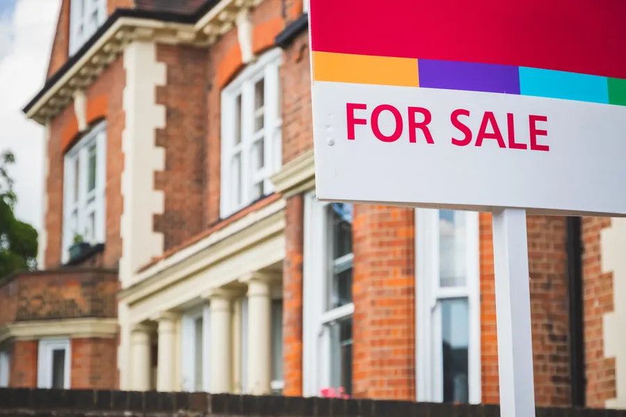 For sale sign in front of a large, red-brick house with white-trimmed windows and columns.