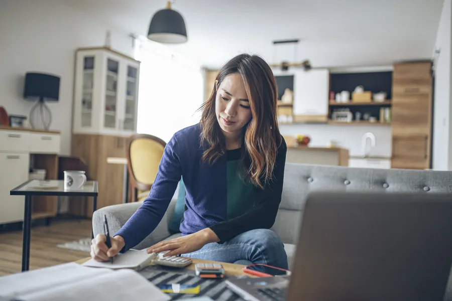 Woman sitting on a couch in a modern living room, writing in a notebook with a laptop and papers on the table.