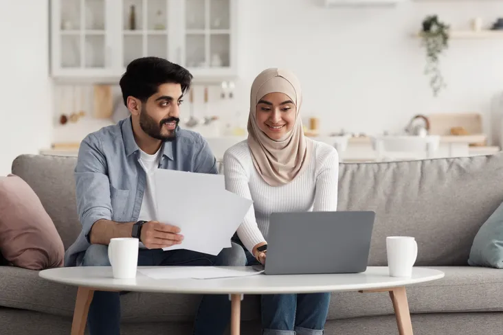 A smiling couple sits on a couch, using a laptop to look at Sharia mortgages and holding papers, with coffee mugs on the table in a bright, modern living room.