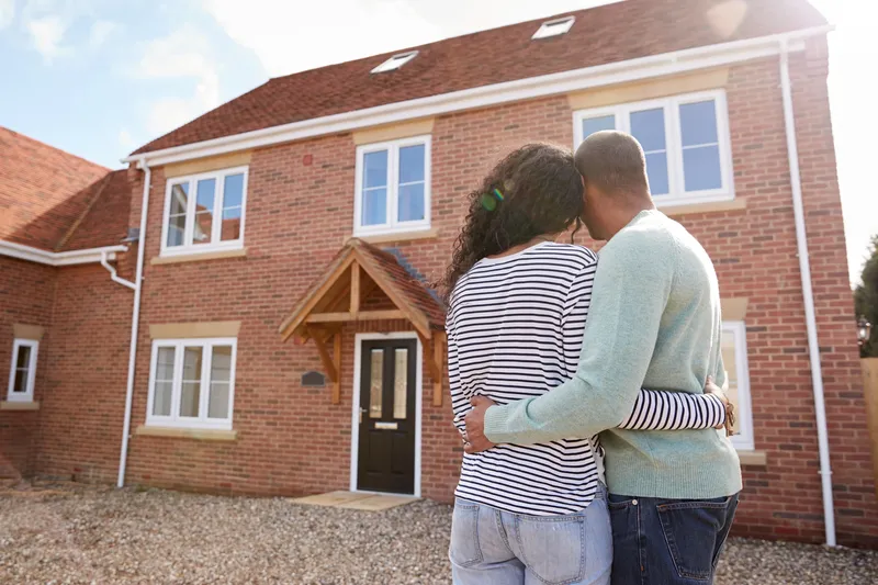 A couple embraces while standing in front of a newly purchased brick house, looking at it with anticipation and happiness as first time buyers.