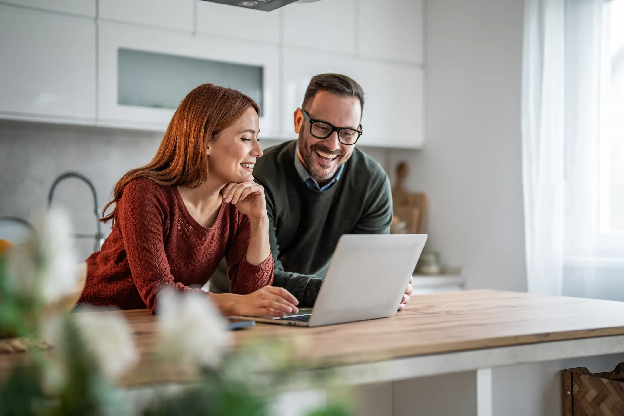 A man and a woman smiling and looking at a laptop on a kitchen counter.
