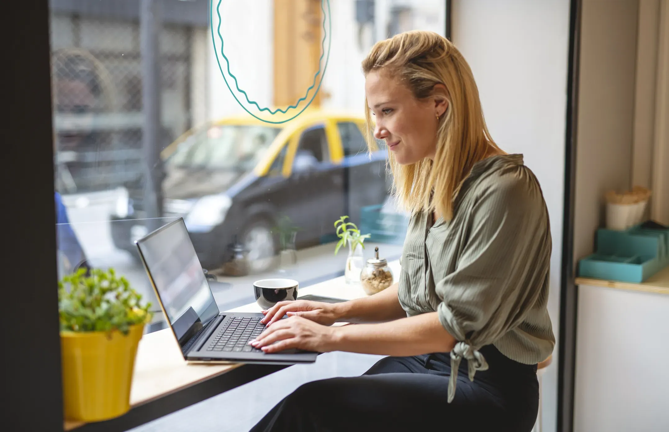 A woman with blond hair works on a laptop at a cafe window seat, with a coffee cup and potted plant nearby. A car is visible through the window.
