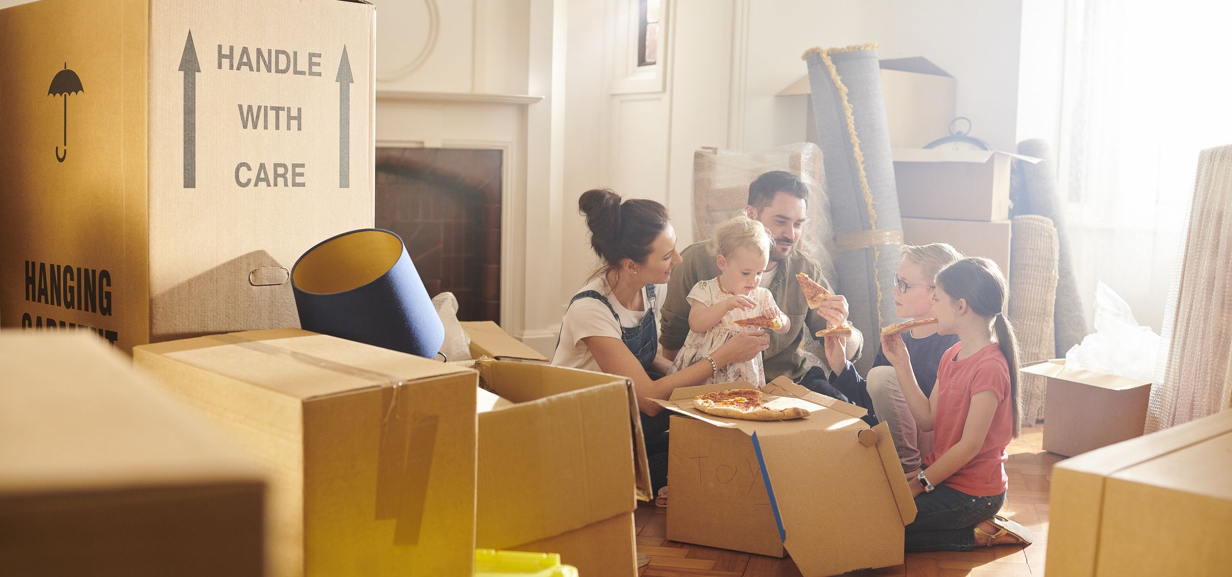 Family sitting on the floor eating pizza amid moving boxes in a sunlit room.