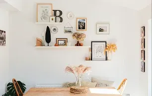 A cozy dining area with a wooden table, dried flowers in a vase, and a gallery wall featuring framed art and decor on white walls.