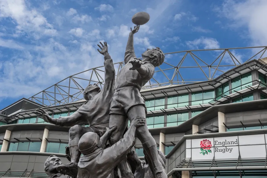 Bronze rugby statue depicting players reaching for a ball, set against Twickenham Stadium with an "England Rugby" sign.