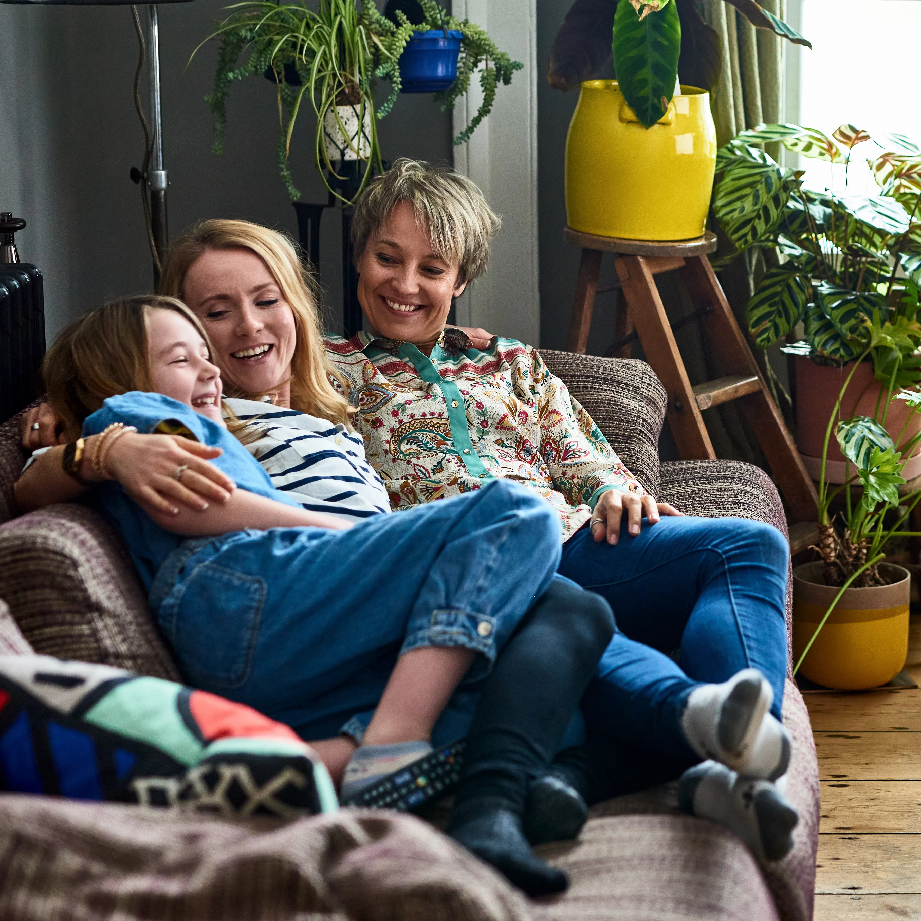 Three people are joyfully sitting on a couch in their new home. A child is cuddled between two adults, laughing. The room is decorated with vibrant plants and a yellow pot. The atmosphere is warm and cozy.
