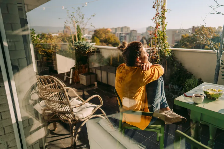 Woman in a yellow shirt sitting on a balcony, gazing at the cityscape. A wicker chair and table with a bowl are nearby.