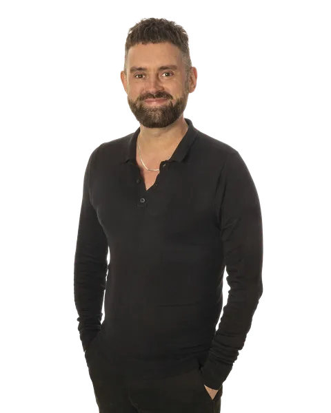 Smiling man with a beard, wearing a black sweater, poses with hands in pockets