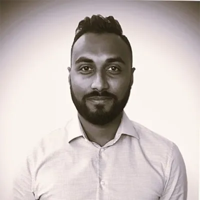 Kavi with a short beard and styled hair, wearing a light-colored button-up shirt, poses against a plain background. Image is in black and white.
