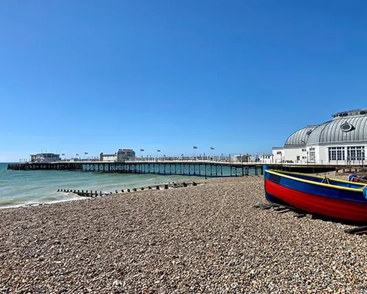 Pebble beach with a colourful boat, Worthing pier extends into the sea, and the glass pavilion building sits under a clear blue sky.