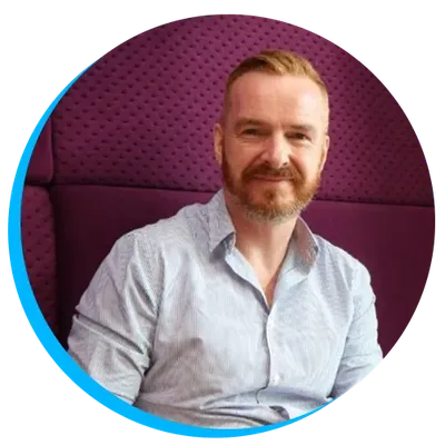 Head shot of Alistair Singer with a beard sitting on a purple chair, wearing a light blue shirt, and smiling at the camera.