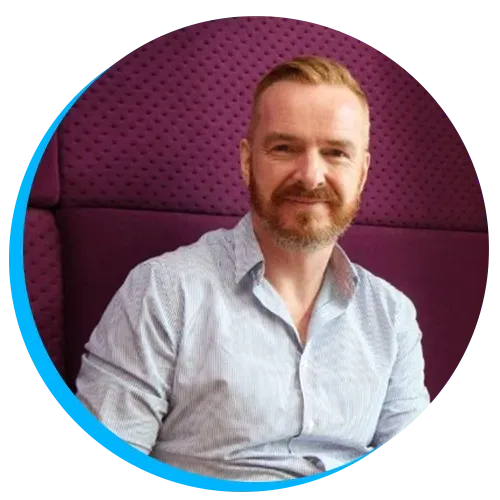 Head shot of Alistair Singer with a beard sitting on a purple chair, wearing a light blue shirt, and smiling at the camera.