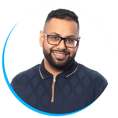 Headshot of Uvais Patel with glasses, beard, and short hair, wearing a dark patterned shirt with a zippered collar, smiling at the camera against a white background.