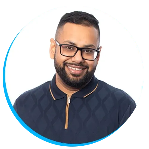 Headshot of Uvais Patel with glasses, beard, and short hair, wearing a dark patterned shirt with a zippered collar, smiling at the camera against a white background.