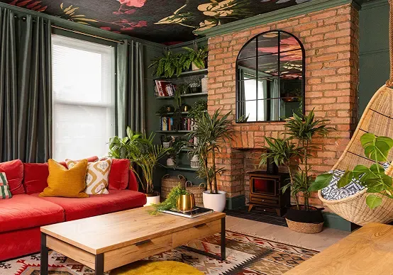 Cozy living room with red sofa, brick fireplace, plants, and hanging chair. Floral ceiling and large mirror add charm. Warm, inviting decor.