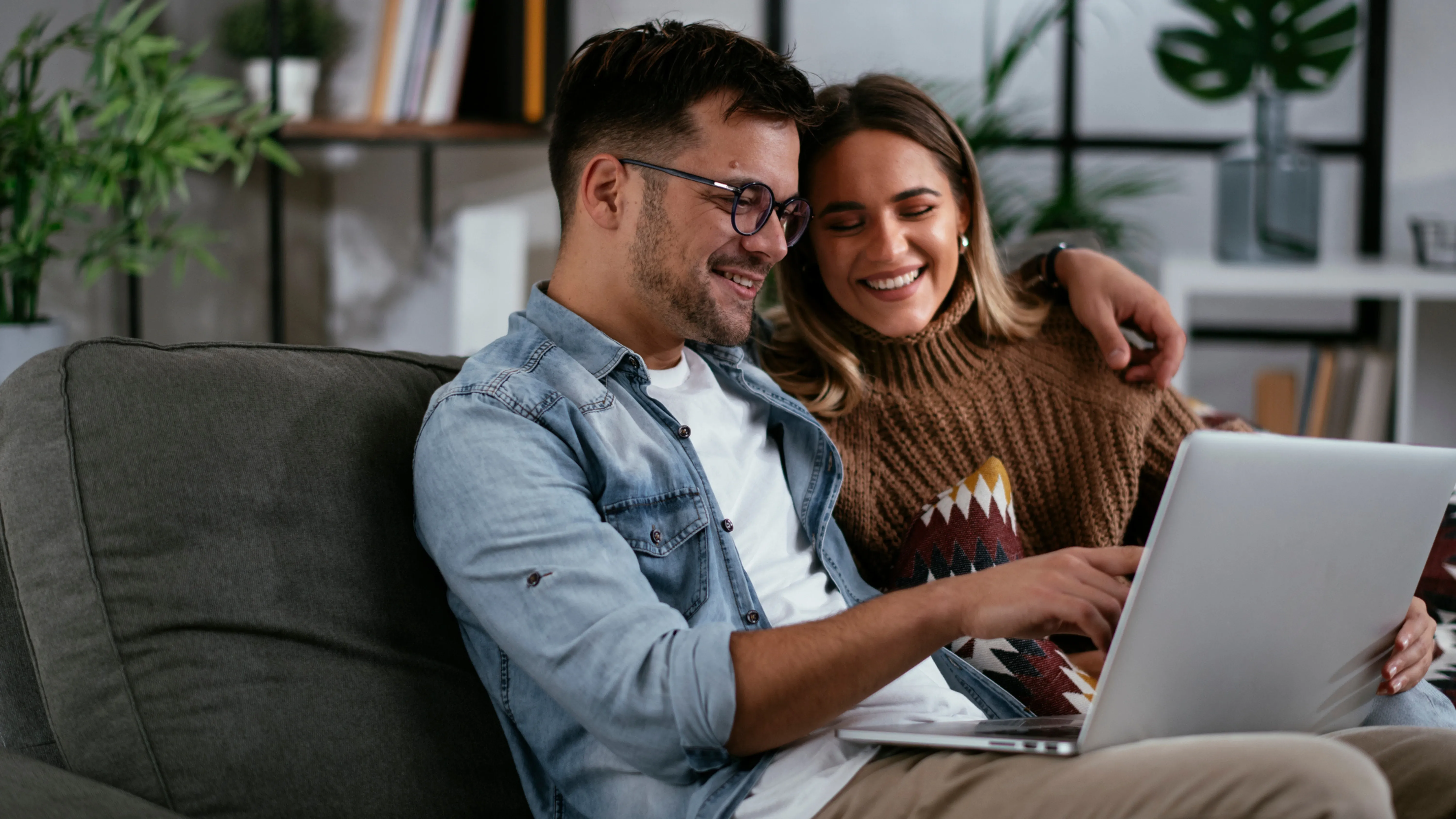 A couple sitting on a couch, smiling and looking at a quote for their limited company from My Home Move Conveyancing on a laptop together in a cozy, plant-filled living room.