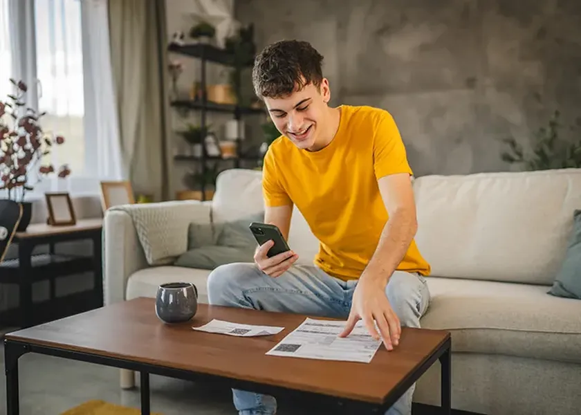 A person in a yellow shirt sits on a couch, smiling at a smartphone, with papers and a mug on the coffee table in front.