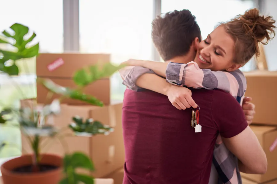 A woman happily hugs a man while holding keys, surrounded by moving boxes and a houseplant, symbolizing a new home.