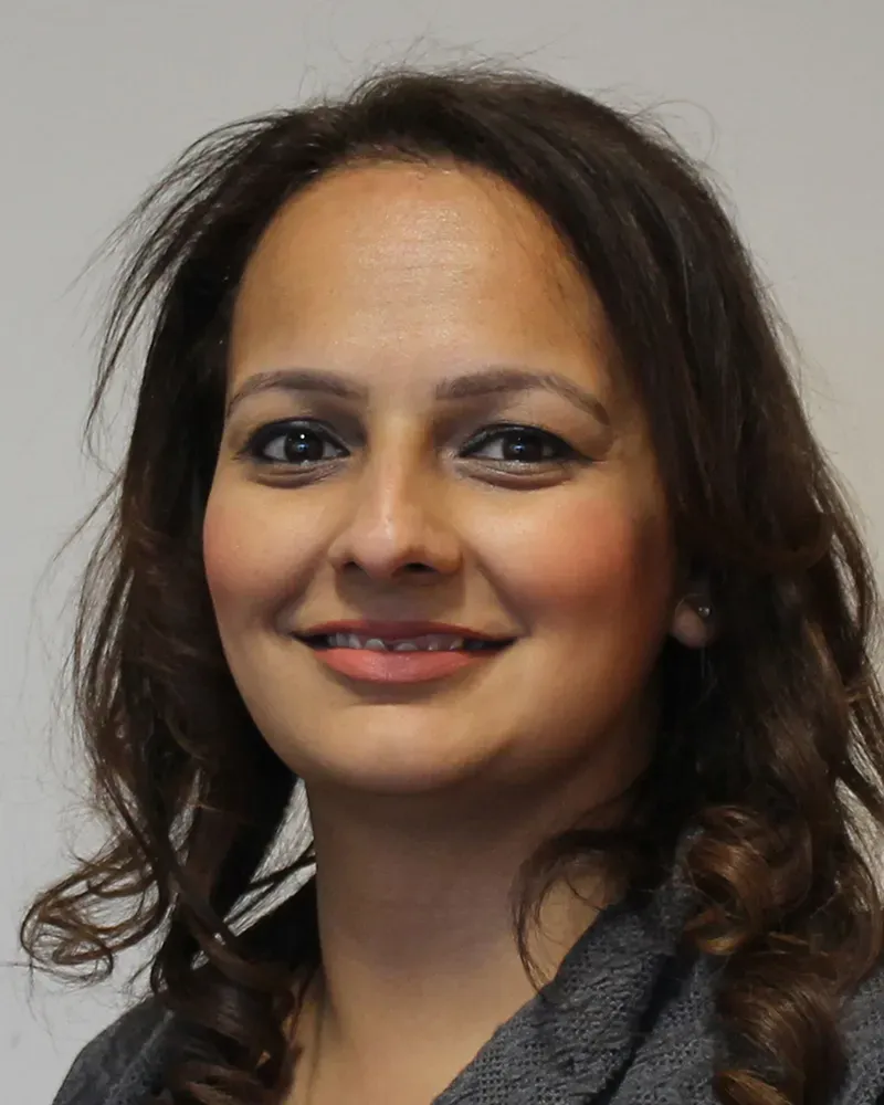 Head and shoulder profile picture of Parminder Phull smiling with long, wavy brown hair, wearing a gray top, against a plain background.