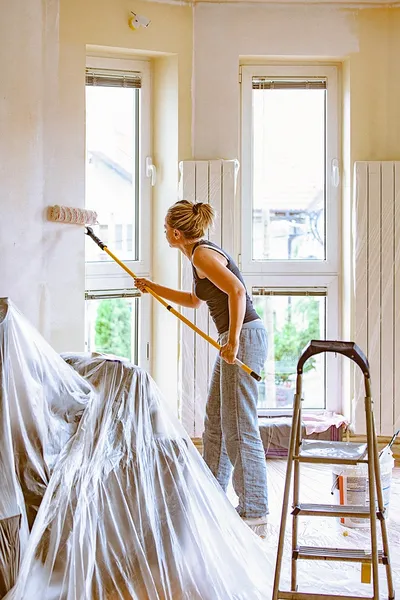 Woman painting a room with a roller, furniture covered in plastic, ladder nearby, natural light from large windows.