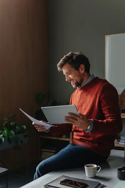 Man in a red sweater sits on a desk, smiling while holding a paper and tablet. A cup of coffee and tablet are in front of him.