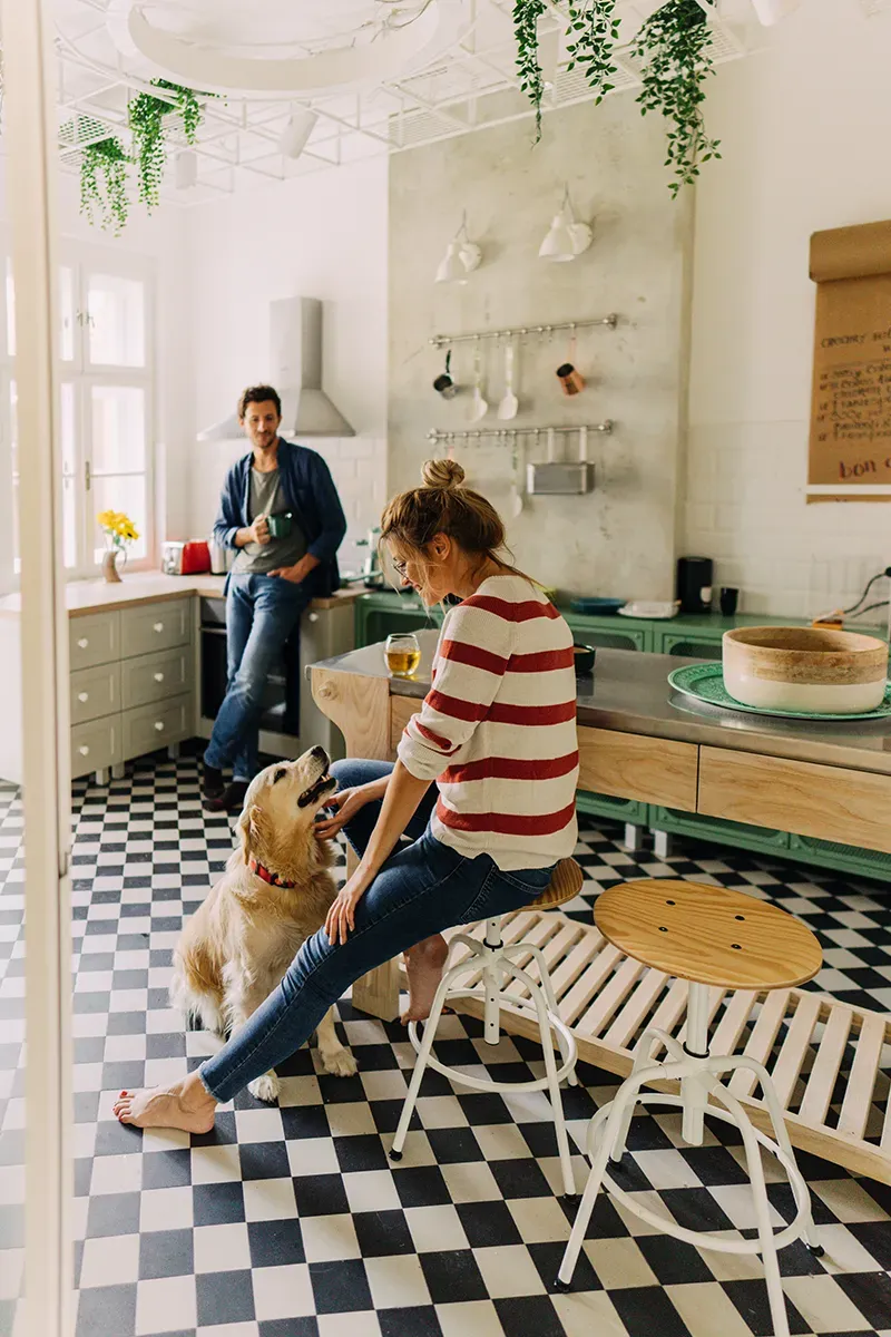 A couple are in their modern country kitchen, he stands by the sink with a cup of coffee and she is sitting by the island stroking their labrador dog.