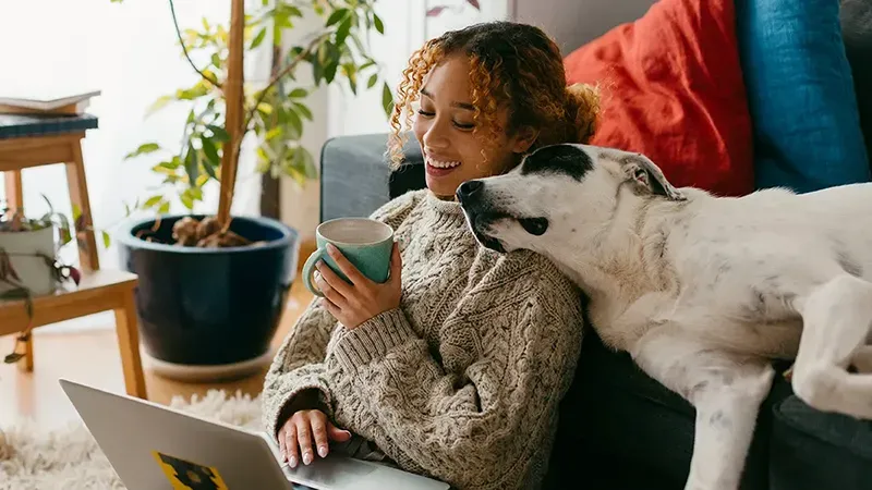 a young lady sitting on the floor of her living room on a soft hug resting against her sofa, where he dog is lying down and resting his head on her shoulder. She is using her laptop to look online at the conveyancing quote for her home move