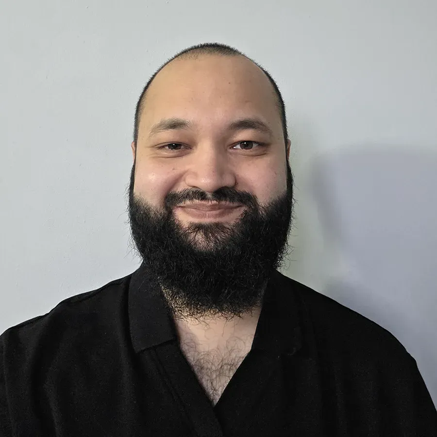Smiling person with a beard and short hair wearing a black shirt, standing against a plain light gray background.