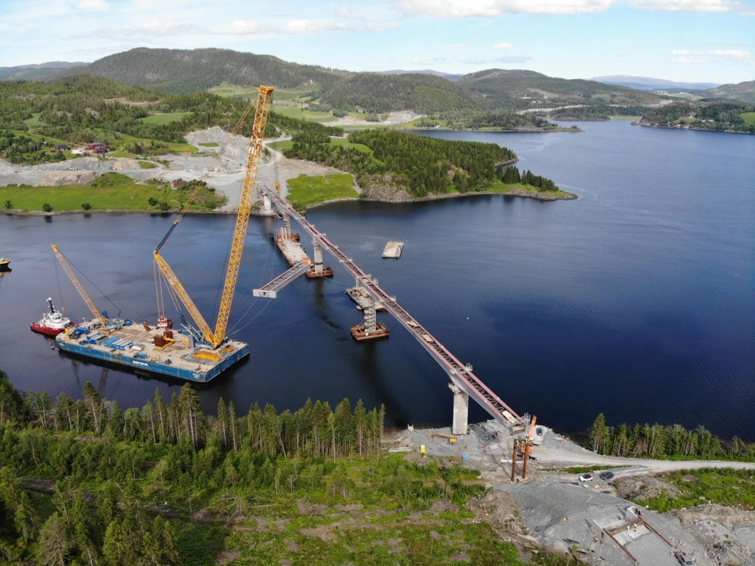 Aerial view of a bridge under construction over a wide river, with cranes and construction barges, surrounded by lush green hills.
