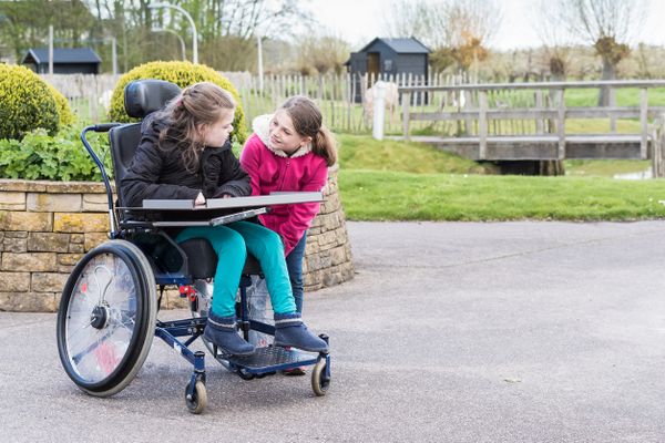 Ein Mädchen im Rollstuhl, gemeinsam mit einem anderen Mädchen ohne Rollstuhl auf einem Schulhof.