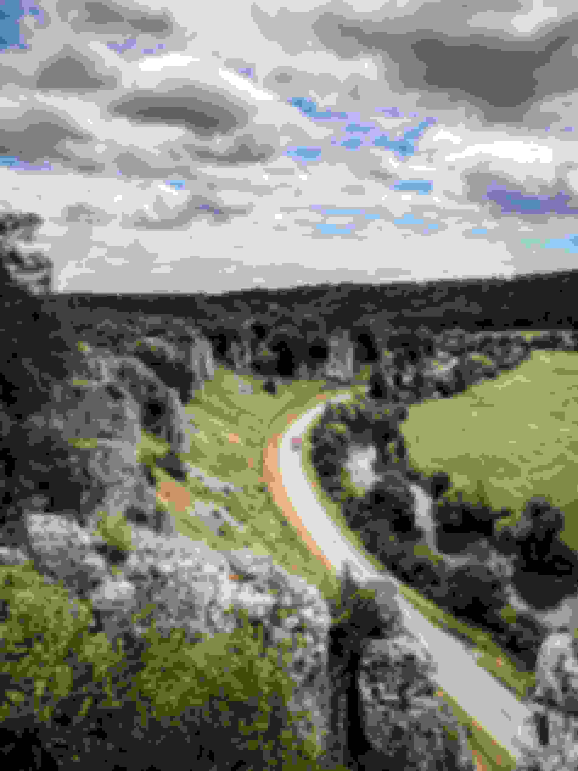 Eine kurvenreiche Straße schlängelt sich durch eine üppige Landschaft mit felsigen Klippen, einem Fluss und weiten grünen Wiesen unter einem teils bewölkten Himmel.