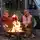 Zwei Männer sitzen vor einem mit Lichterketten geschmückten Campervan an einem Lagerfeuer und stoßen mit Flaschen aufeinander an.