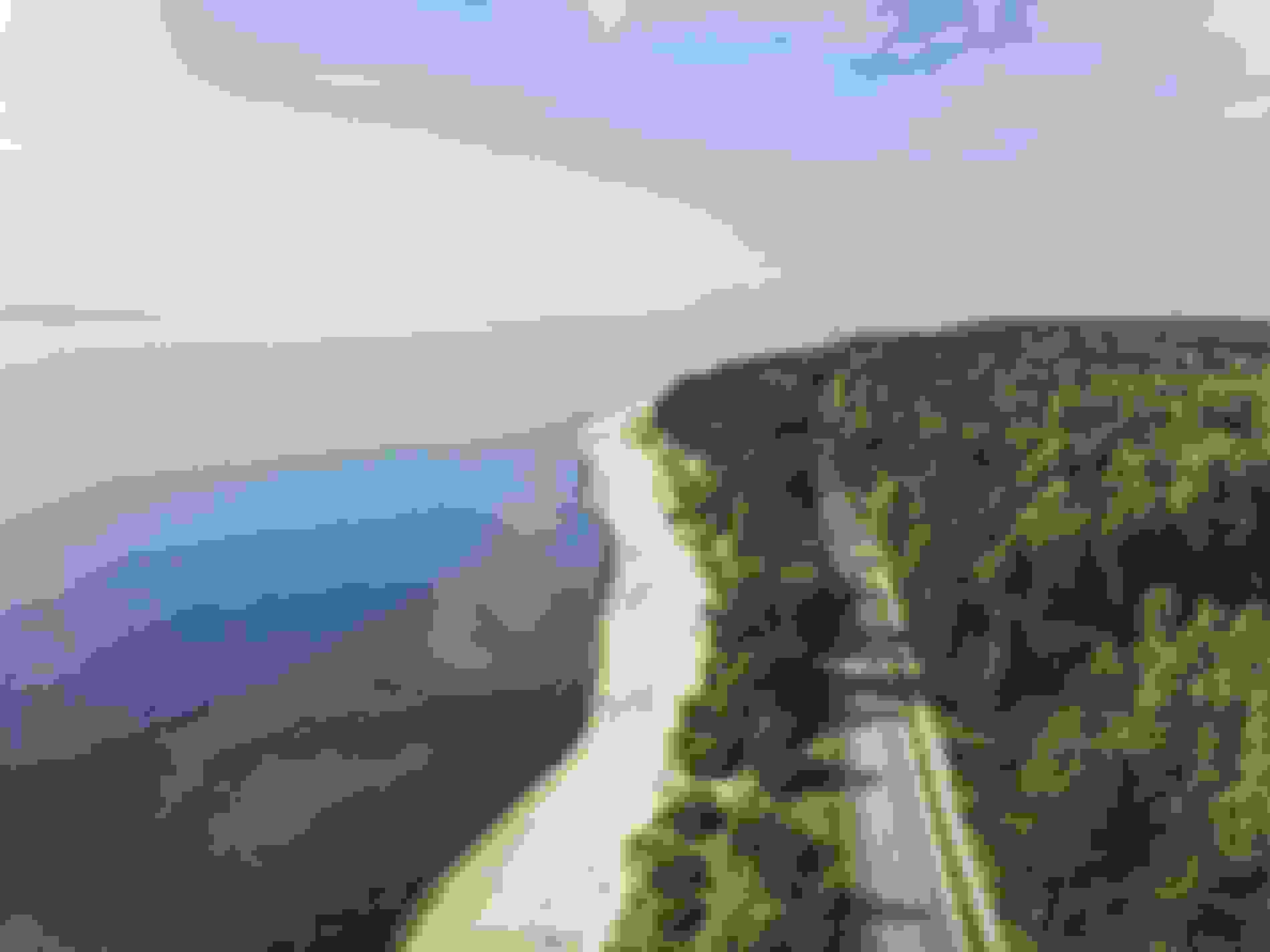 Luftaufnahme einer ruhigen Küstenlandschaft mit einem Sandstrand, üppig grünem Wald und einer Straße, die parallel zur Küste verläuft – unter einem klaren blauen Himmel.
