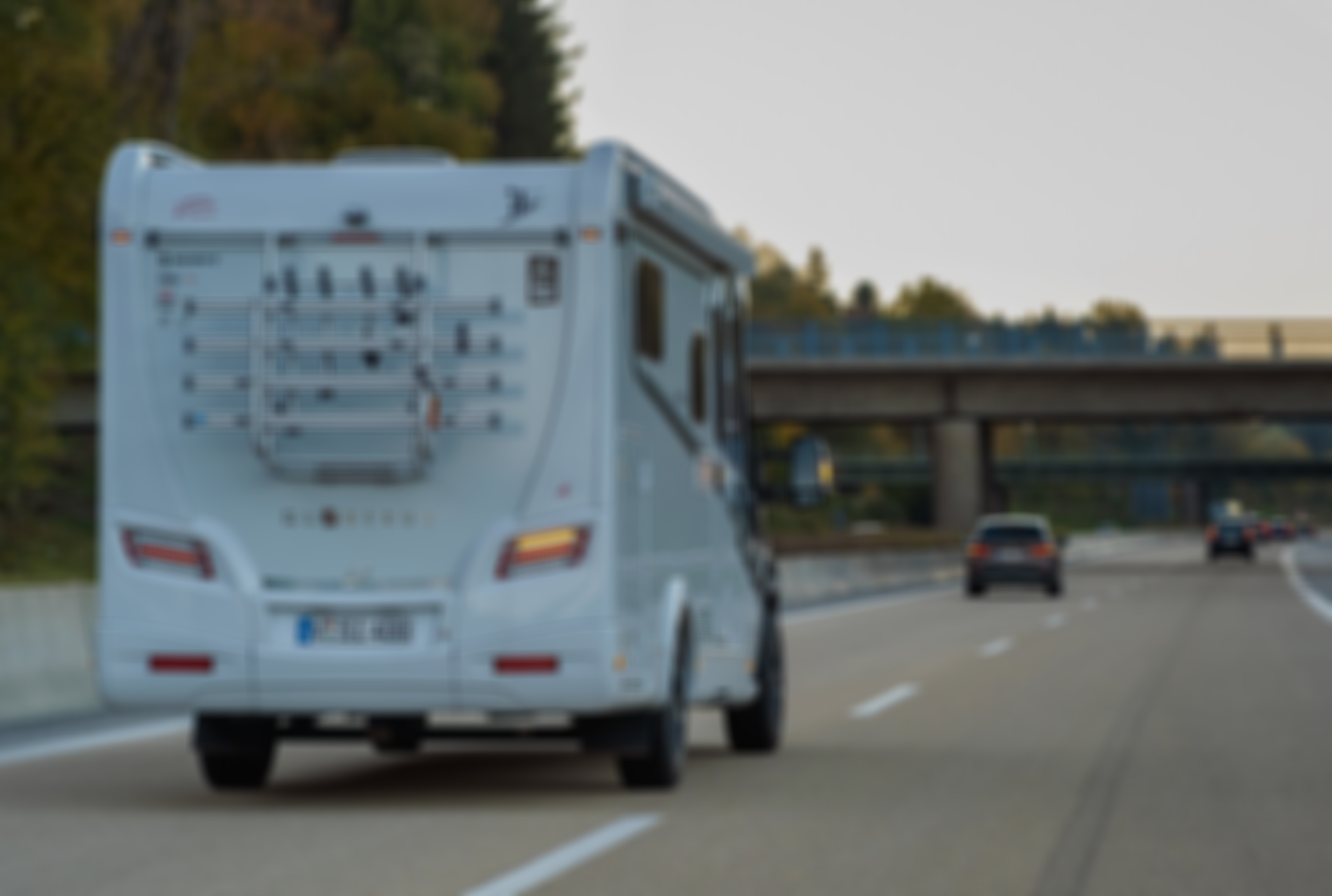 Ein weißes Wohnmobil fährt auf einer Autobahn, hinten ist ein Fahrradträger montiert. Mehrere Autos sind voraus, im Hintergrund ist eine Brücke zu sehen.