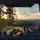 Blick aus dem Inneren eines Campers: Ein schlafender Hund und zwei zugedeckte Personen genießen den Ausblick auf eine bewaldete Landschaft bei Sonnenuntergang.
