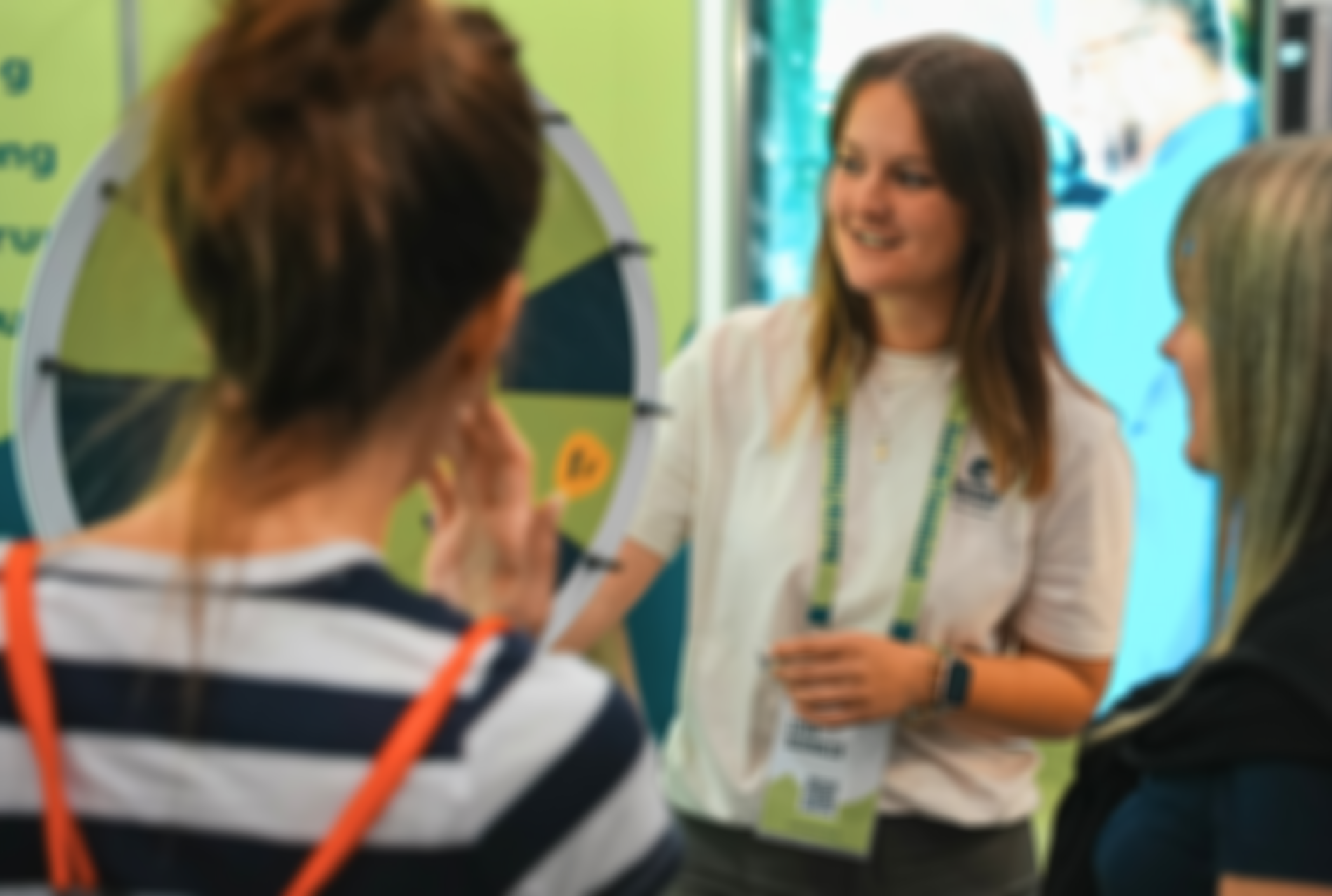 Eine Frau im weißen Shirt mit Lanyard spricht auf einer Veranstaltung mit zwei Besucherinnen neben einem interaktiven Glücksrad.