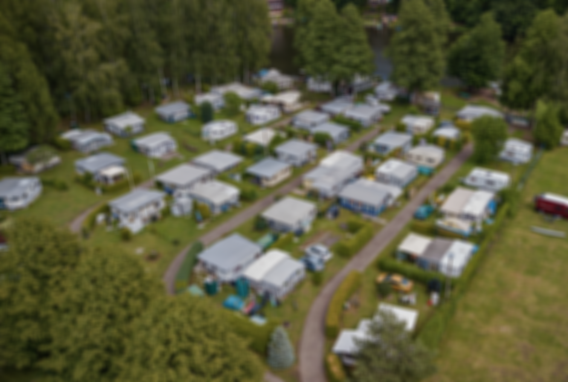 Luftaufnahme eines Campingplatzes mit Reihen von Wohnwagen und Zelten, umgeben von Bäumen, in der Nähe eines Flusses. Wege verbinden die einzelnen Stellplätze.
