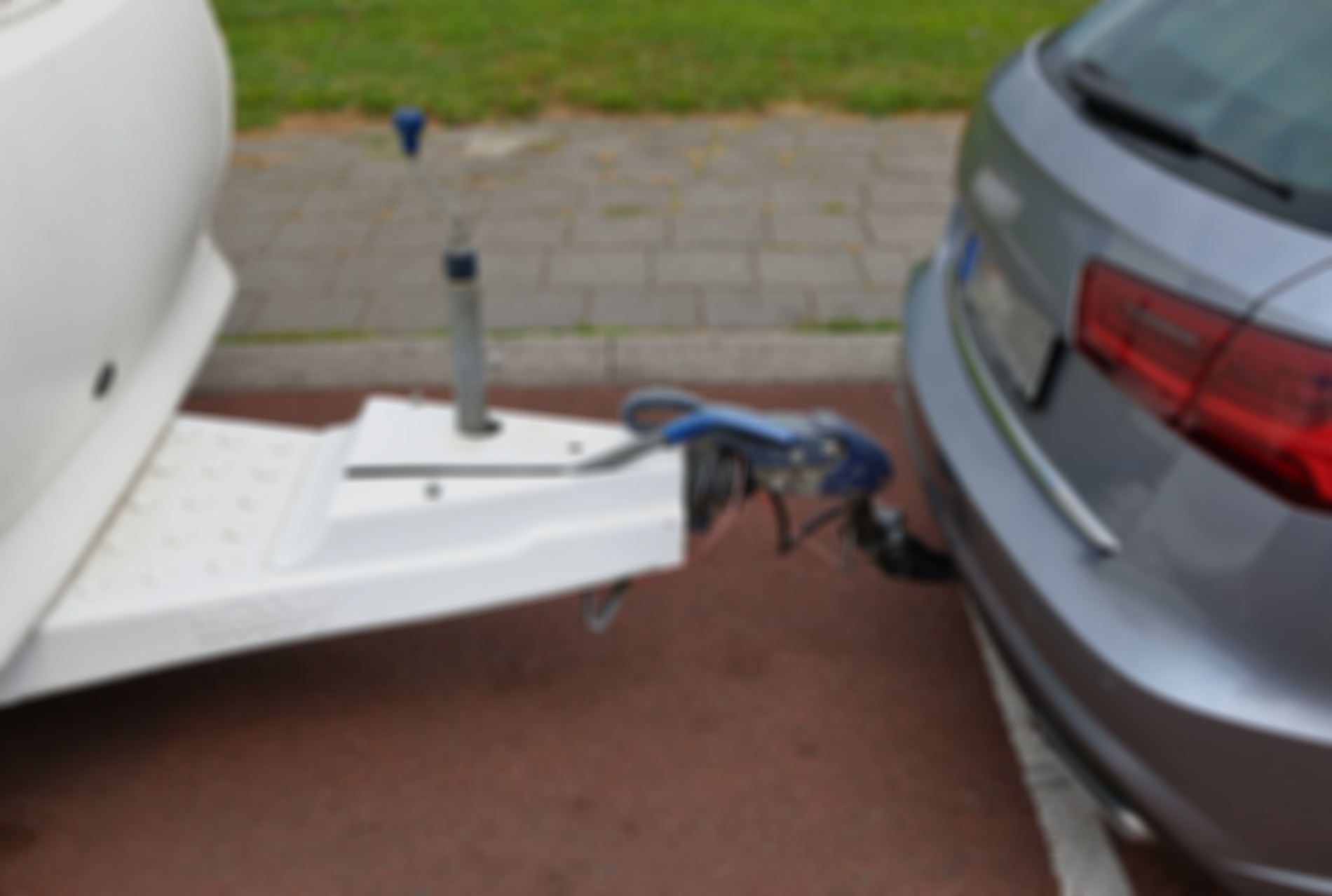 Ein Auto ist an einen weißen Anhänger angekuppelt und steht auf einer asphaltierten Straße, im Hintergrund sind Gras und ein Gehweg zu sehen.