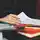 Eine Person hält einen Stift in der Hand und prüft an einem Schreibtisch einen Stapel Dokumente. Daneben liegen ein orangefarbenes Notizbuch, im Hintergrund ist eine unscharfe Umgebung zu sehen.