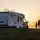 Wohnmobil auf einem grasbewachsenen Hügel bei Sonnenuntergang, daneben stehen zwei Personen und blicken auf den malerischen Horizont.