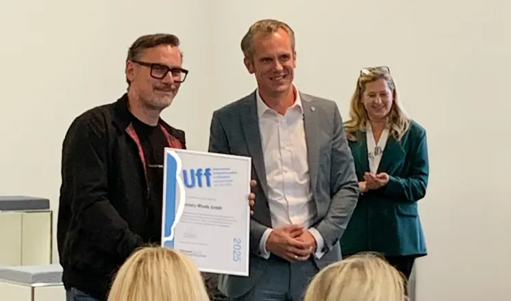 Two men stand at the front of a room during an award ceremony. One of them holds a framed certificate labeled ‘UFF’ while both smile toward the audience. A woman in a green blazer stands in the background applauding. Several people are seated in the foreground, watching the presentation.